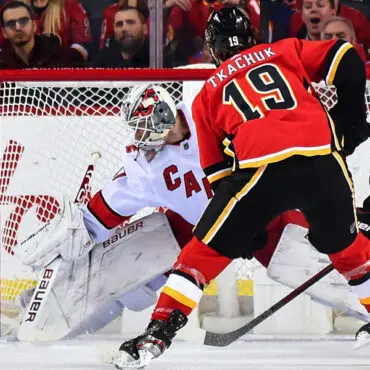 Hockey player in red jersey preparing to score against goalie in white gear.