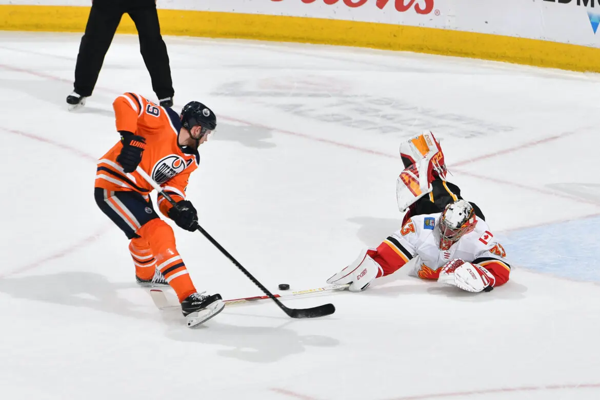 Hockey player in orange shoots the puck past a diving goalie in white and red.