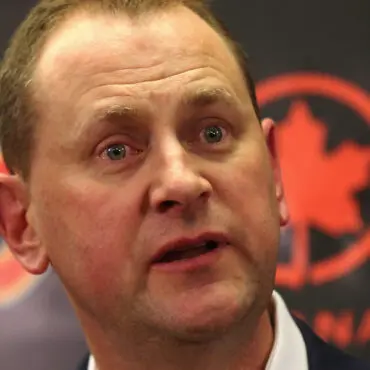 A man speaking with a serious expression in front of a black and red background.