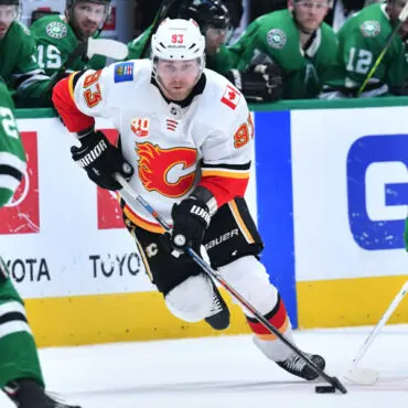 Hockey player in white and red jersey skating with puck on ice rink.