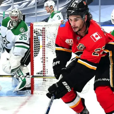 Hockey player in red and black jersey skating with puck near goal.