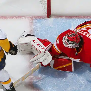 Hockey goalie in red gear making a save against an opponent.