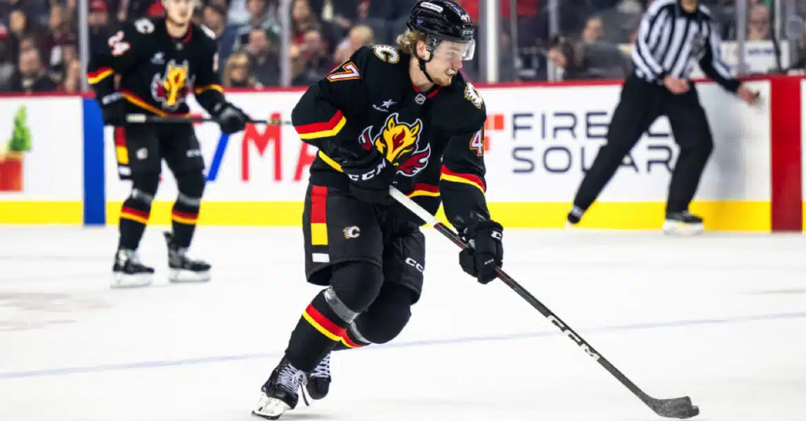 Hockey player in black and red uniform skating with puck on ice rink.