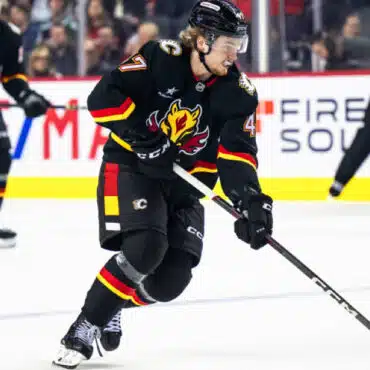 Hockey player in black and red uniform skating with puck on ice rink.
