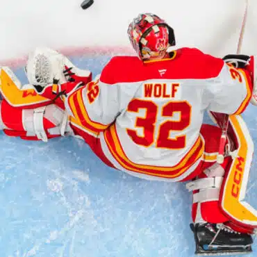 Hockey goalie in red and white gear sprawled on ice during a game.