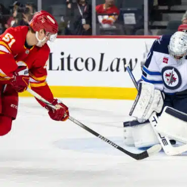 A hockey player in red shoots the puck past a goalie in white gear.